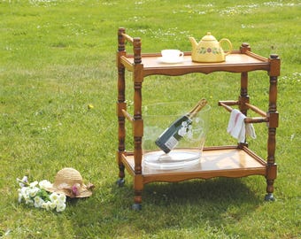 Puede incluir: Un carrito de té de madera de dos niveles con una tetera amarilla, una taza de té y una botella de champán en una cubitera. Un sombrero de paja con flores descansa sobre la hierba. El carrito tiene un toallero.