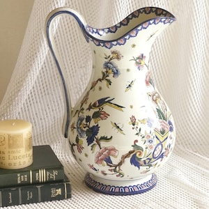 May include: A large, white ceramic jug with a curved handle and a wide spout. The jug is decorated with colourful floral and bird motifs, with blue and pink accents. A candle and books are in the foreground.