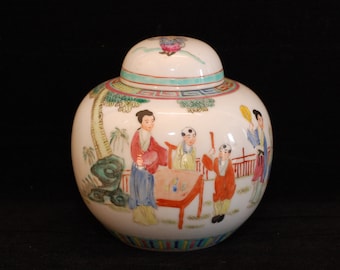 Puede incluir: Un tarro de porcelana blanca con tapa, decorado con una escena colorida de personas en un jardín. El tarro tiene un borde verde, azul y rojo alrededor de la base.