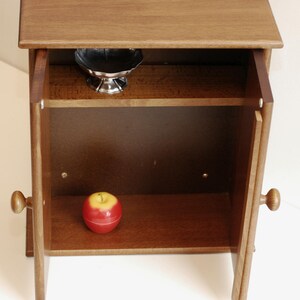 May include: A small wooden cabinet with two doors, both open. The cabinet has one shelf inside, and a red apple is sitting on the bottom shelf. A small silver bowl is on the top shelf.