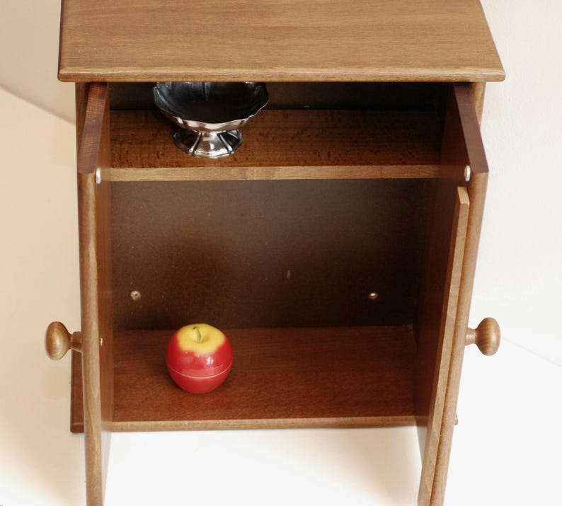 May include: A small wooden cabinet with two doors, both open. The cabinet has one shelf inside, and a red apple is sitting on the bottom shelf. A small silver bowl is on the top shelf.