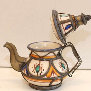 Peut inclure: Une petite théière en céramique blanche avec un motif floral bleu, vert et orange. La théière a un couvercle et une poignée en métal avec des détails complexes. La théière est décorée d'une bordure en métal autour du bord et de la base.