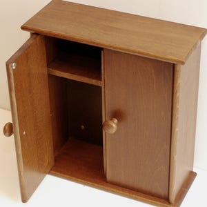 May include: A small wooden cabinet with a single door that is open. The cabinet has a shelf inside and a knob on the door. The cabinet is a light brown color.