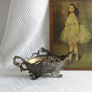 Op de afbeelding: Een antieke, sierlijke metalen kom met een bronzen afwerking, met ingewikkeld krulwerk en handvatten. De kom staat tegen een achtergrond van een vintage schilderij van een jong meisje in een jurk en een witte, getextureerde stof.