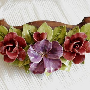 May include: A ceramic flower arrangement with three red roses and a purple flower, all with green leaves. The flowers are attached to a brown wooden base.