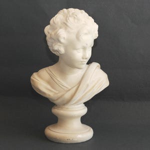 May include: A cream-coloured bust of a young person with curly hair and a draped garment. The sculpture is mounted on a pedestal base. The bust is a classical art piece, likely made of plaster or a similar material.