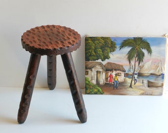 May include: A wooden three-legged stool with a round, textured top. The stool is brown and has a rustic appearance. A painting of a tropical scene with palm trees, houses, and people is in the background.