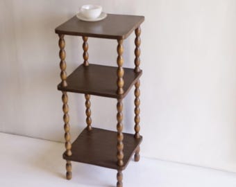 May include: A three-tiered dark brown wooden stand featuring square shelves and four vertical supports with a decorative turned spindle design. A small white ceramic teacup and saucer are placed on the top shelf of the furniture piece.