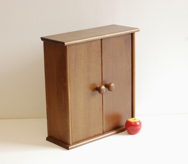 May include: A small wooden cabinet with two doors and round knobs. The cabinet is a light brown color and has a simple design.