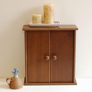 May include: A small wooden cabinet with two doors and brown knobs. The cabinet is sitting on a white surface. There are two candles and a tray on top of the cabinet.