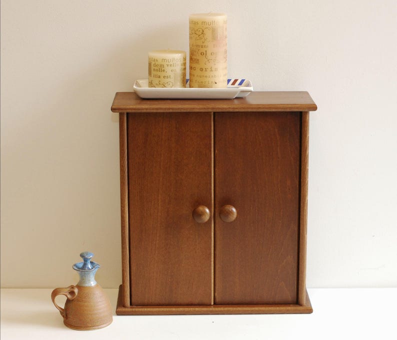 May include: A small wooden cabinet with two doors and brown knobs. The cabinet is sitting on a white surface. There are two candles and a tray on top of the cabinet.