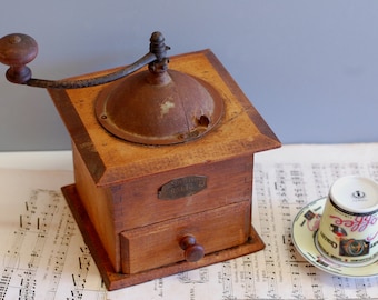 May include: Antique wooden coffee grinder with a metal crank and a drawer for storing coffee grounds. The grinder has a rusty metal top and a label that reads "Cafe Moulu." The grinder is sitting on a sheet of music.