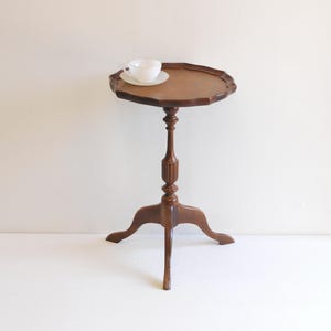 Puede incluir: Una pequeña mesa auxiliar antigua de madera marrón con una encimera con borde festoneado. La mesa tiene una base de pedestal con tres patas. Una taza de té y un platillo blancos están sobre la encimera. La mesa mide unos 71 cm de alto.