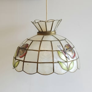 Op de afbeelding: Een witte, schelpvormige hanglamp met een gouden rand en vlinderontwerpen. De lamp heeft een rasterpatroon en een gegolfde rand.