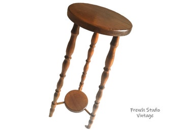 May include: A wooden plant stand with a round top and a lower shelf. The stand has three legs with turned spindles.