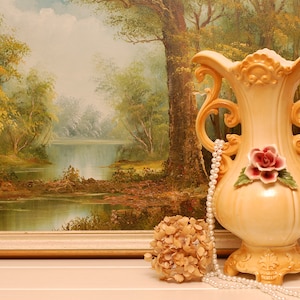 Peut inclure: Un vase en céramique blanche avec des détails ornés et un motif floral. Le vase est décoré d'une rose rose et de feuilles vertes. Le vase est posé sur une table devant une peinture de paysage.