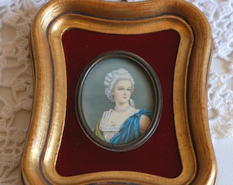 May include: A framed miniature portrait of a woman with a white powdered wig and a blue and white dress. The frame is gold with a red velvet mat.