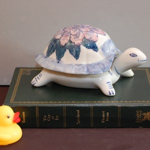Puede incluir: Una figura de tortuga de cerámica con cuerpo blanco y caparazón decorado con flores rosas y hojas azules. La tortuga está sobre un libro verde junto a un patito de goma amarillo. La figura es un objeto decorativo.