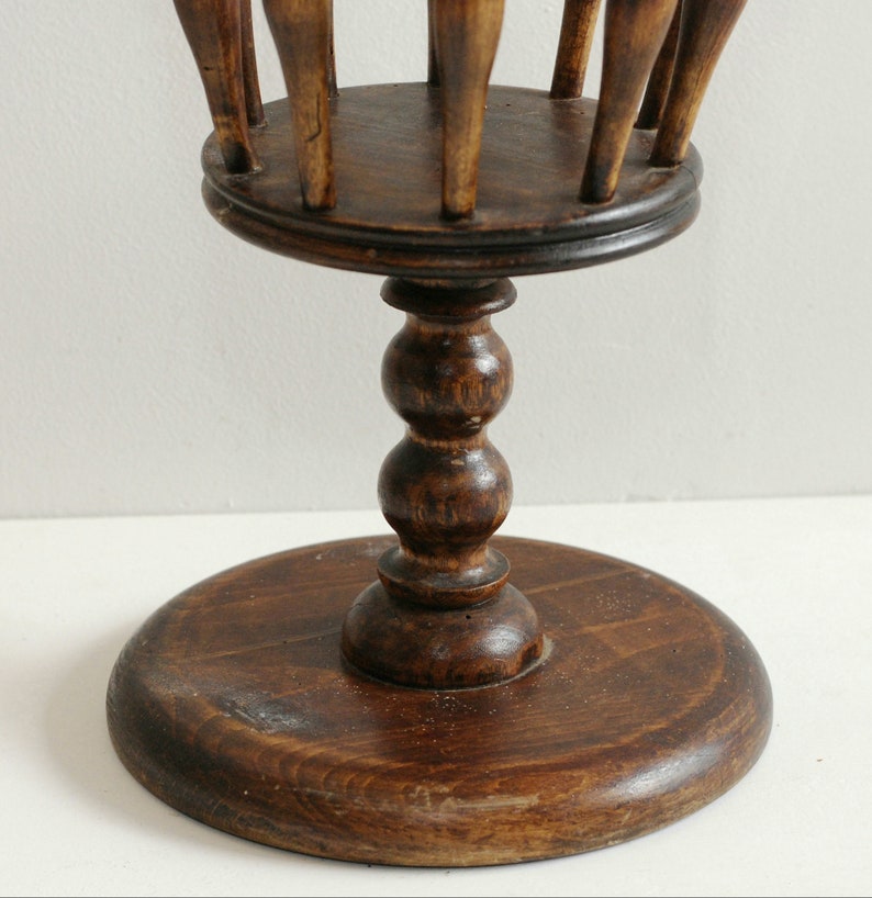 May include: A wooden stand with a round base and a pedestal with a decorative ball design. The top of the stand has a circular opening with wooden dowels sticking up.
