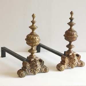 May include: A pair of antique brass and black iron fire dogs. The brass elements have an ornate, detailed design with a classical motif. The fire dogs have a decorative top and a base with a lion's head. The black iron supports the brass elements.