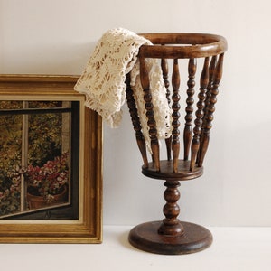 May include: A wooden basket with a turned spindle design, standing on a pedestal base. The basket is empty, but a white crocheted doily is draped over the top.