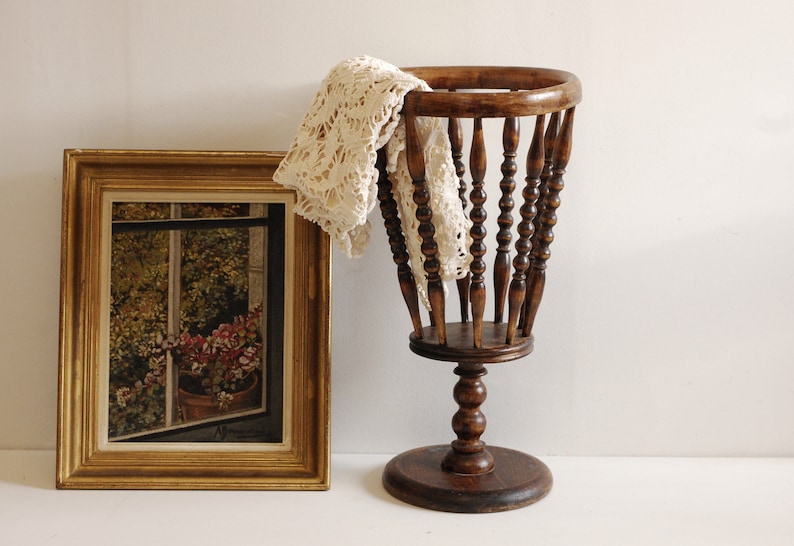 May include: A wooden basket with a turned spindle design, standing on a pedestal base. The basket is empty, but a white crocheted doily is draped over the top.