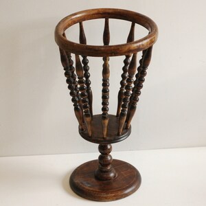 May include: A wooden umbrella stand with a round top and a base. The stand is made of dark brown wood and has a decorative design of small beads.