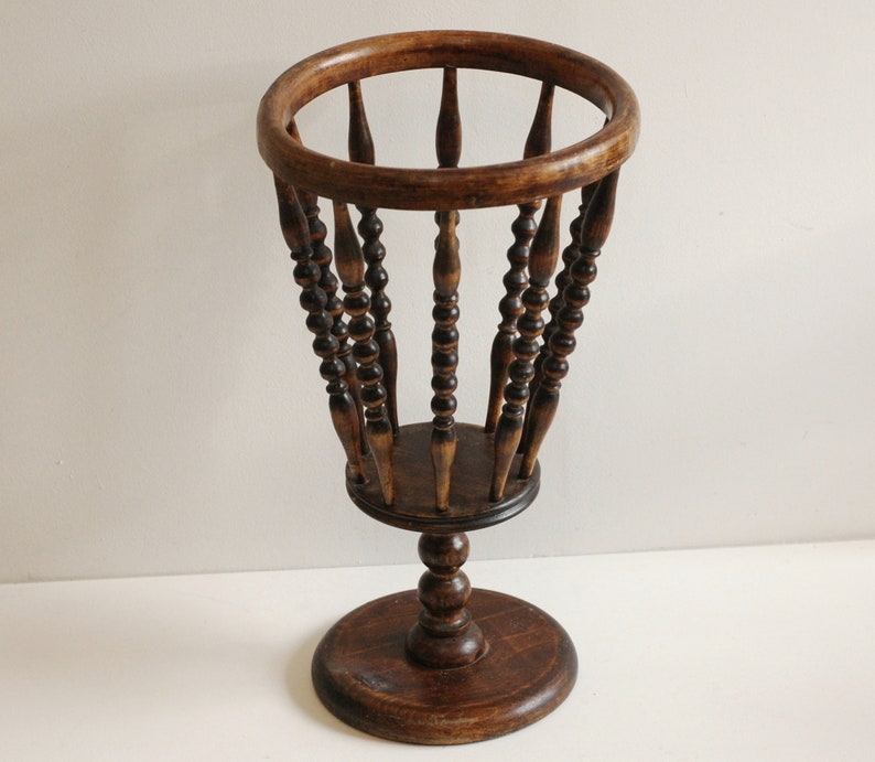 May include: A wooden umbrella stand with a round top and a base. The stand is made of dark brown wood and has a decorative design of small beads.