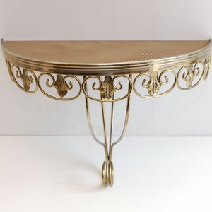 Puede incluir: Una estantería de pared de metal dorado con una parte superior semicircular y adornos de volutas. La estantería tiene una parte superior de malla metálica.