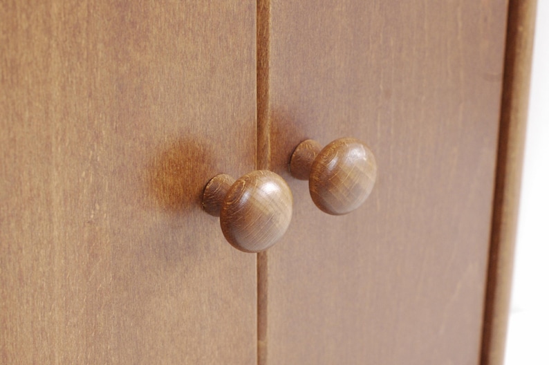 May include: Two wooden cabinet knobs with a rounded, mushroom-shaped design. The knobs are a light brown color and are mounted on a wooden cabinet door.