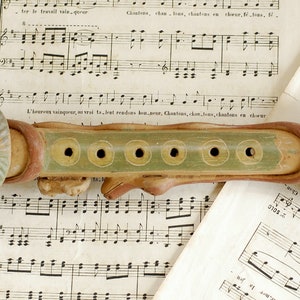 Può includere: Un flauto in ceramica con un designo verde, marrone e giallo. Il flauto ha cinque fori ed è decorato con una testa di animale stilizzata a un'estremità.