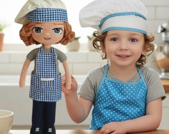 May include: A crochet doll with auburn hair wearing a chef hat and a blue and white checkered apron over a gray shirt and dark pants, standing next to a child wearing a white chef hat and a blue polka dot apron in a kitchen setting.