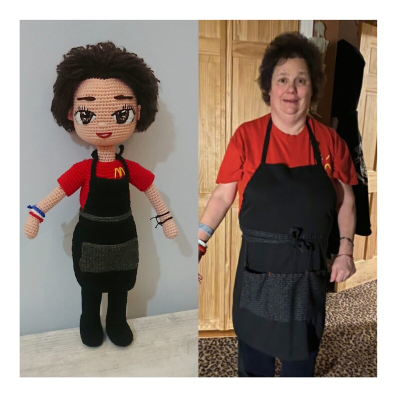 May include: A crocheted doll wearing a red shirt and a black apron with a pocket. The doll has brown hair and large brown eyes. The doll is standing on a wooden floor.