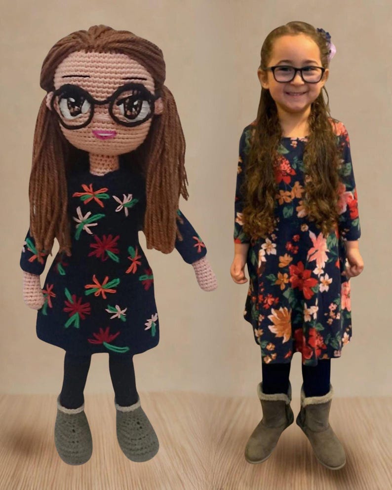 Personalized look-alike crochet amigurumi doll standing next to a smiling girl in a matching dark floral dress. Both wear round glasses and grey boots. The custom handmade amigurumi doll is a detailed replica of the girl based on a photo, making it a unique personalized gift for children or as a custom portrait art piece. White background and light wood parquet floor.