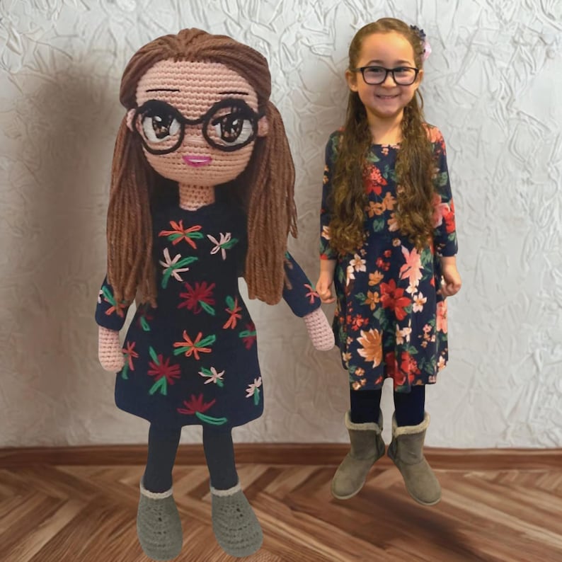 Personalized look-alike crochet amigurumi doll standing next to a smiling girl in a matching dark floral dress. Both wear round glasses and grey boots. The custom handmade amigurumi doll is a detailed replica of the girl based on a photo, making it a unique personalized gift for children or as a custom portrait art piece. White background and light wood parquet floor.