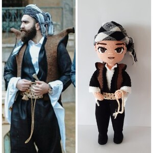 May include: A crocheted doll wearing a black and white patterned hat, a brown vest, and black pants with a white belt. The doll is holding a rope in its right hand.