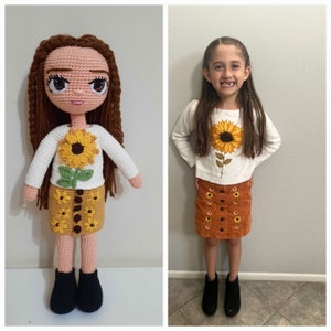 May include: A crocheted doll wearing a white sweater with a sunflower design and a brown corduroy skirt with sunflower embroidery. The doll has brown hair and large brown eyes. The doll is standing next to a young girl wearing a similar outfit.