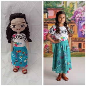 Puede incluir: Una mu&ntilde;eca de crochet que lleva una blusa blanca con bordados negros y una falda turquesa con bordados coloridos. La mu&ntilde;eca tiene el pelo casta&ntilde;o y los ojos marrones. La mu&ntilde;eca est&aacute; de pie sobre una superficie blanca.