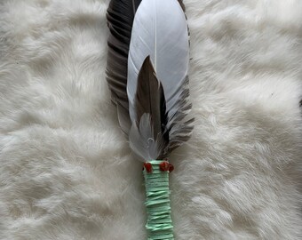 May include: A handcrafted feather fan with a light green wrapped handle. The fan features a mix of black, white, and brown feathers, and is adorned with small red beads. The feathers are arranged in a layered design.