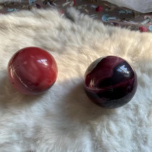 Könnte beinhalten: Zwei polierte, kugelförmige Steine. Einer ist tiefrot mit hellerer roter Marmorierung, der andere ist dunkelviolett mit schwarzen und weißen Wirbeln. Die Steine liegen auf einer weißen, flauschigen Oberfläche.
