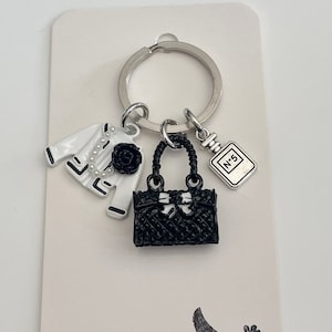Könnte beinhalten: Ein silberner Schlüsselanhänger mit einem schwarzen Handtaschenanhänger, einem weißen Jackenanhänger und einem kleinen Parfümflakonanhänger. Die Anhänger sind an einem silbernen Ring befestigt.