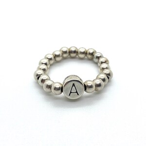 Könnte beinhalten: Ein silberner Ring mit einem Perlenband und einem runden Anhänger mit dem eingravierten Buchstaben 'A'.