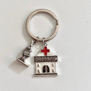 Puede incluir: Llavero de plata con un dije de edificio blanco que dice "HOSPITAL" y una cruz roja en la parte superior. Un dije de microscopio plateado cuelga del dije del edificio.