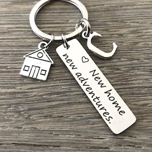 Può includere: Portachiavi d'argento con un ciondolo a forma di casa, un ciondolo a forma di cuore e un'etichetta che recita "Nuova casa, nuove avventure."
