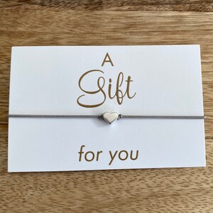 Könnte beinhalten: Eine weiße Karte mit den Worten "A Gift for you" in goldener Schrift. Ein graues Armband aus Schnur mit einem silbernen Herz-Anhänger ist an der Karte befestigt.
