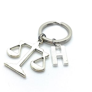 Op de afbeelding: Zilveren sleutelhanger met een weegschaal-bedel en een letter H-bedel.