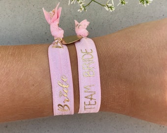 Op de afbeelding: Twee roze elastische armbandjes met gouden letters. Het ene armbandje zegt "Bride" en het andere zegt "Team Bride".