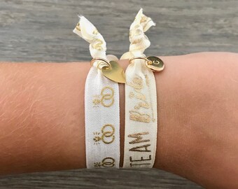Op de afbeelding: Twee ivoorkleurige haarbandarmbanden met gouden tekst en bedels. Eén zegt "Team Bride" en de andere heeft trouwringafbeeldingen. Eén heeft een gouden hartbedel, de andere een gouden initiaalbedel.