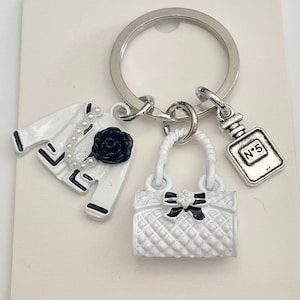Könnte beinhalten: Weißer Schlüsselanhänger mit einer weißen Handtasche, einer weißen Jacke und einem kleinen Parfümflakon-Anhänger. Der Schlüsselanhänger hat einen silbernen Ring und eine schwarze Schleife an der Handtasche.