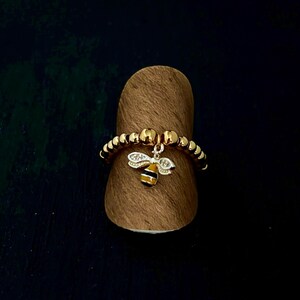 Op de afbeelding: Gouden kralenring met een bijenbedel. De ring heeft kleine, ronde gouden kralen en een bungelende bijenhangertje met witte kristallen accenten en zwart-gele strepen. De ring wordt gepresenteerd op een bruine, cilindrische standaard.